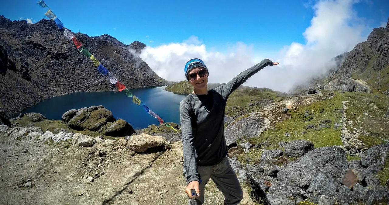 A trekker with arms out beside a turquoise lake ringed by prayer flags and rock in the Langtang hills