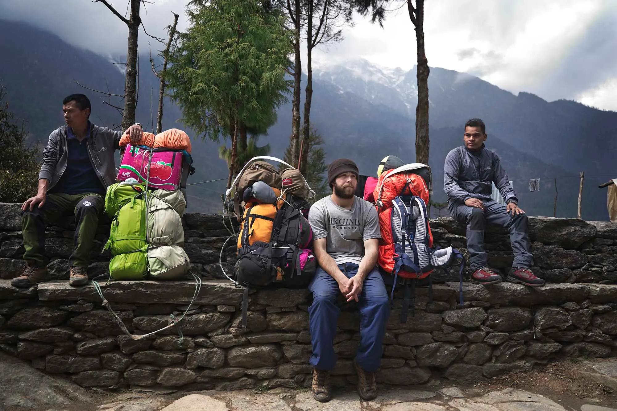 Porters resting on a stone wall beside their loads under tall trees on a Nepal trail