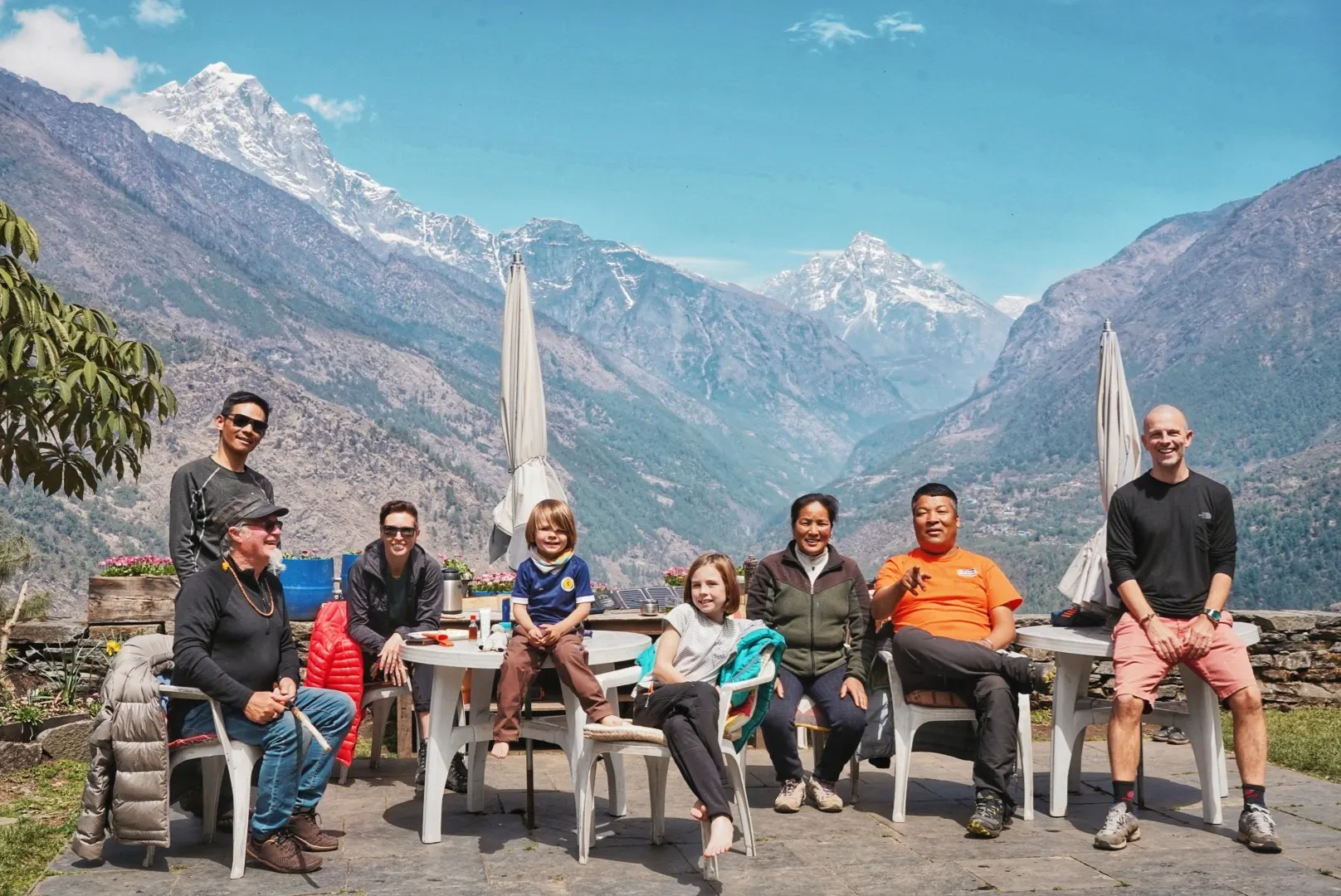 A group of trekkers resting at an outdoor teahouse table with Khumbu peaks behind